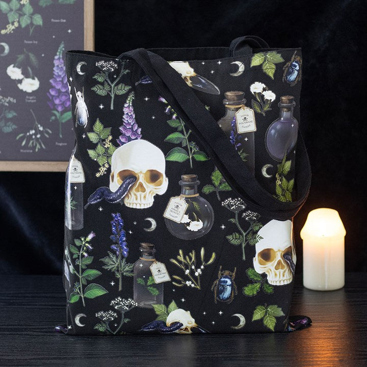 A gothic botanical tote bag adorned with skulls, potion bottles, snakes, purple flowers, green leaves, and crescent moons rests on a dark surface while a lit candle glows in the background.