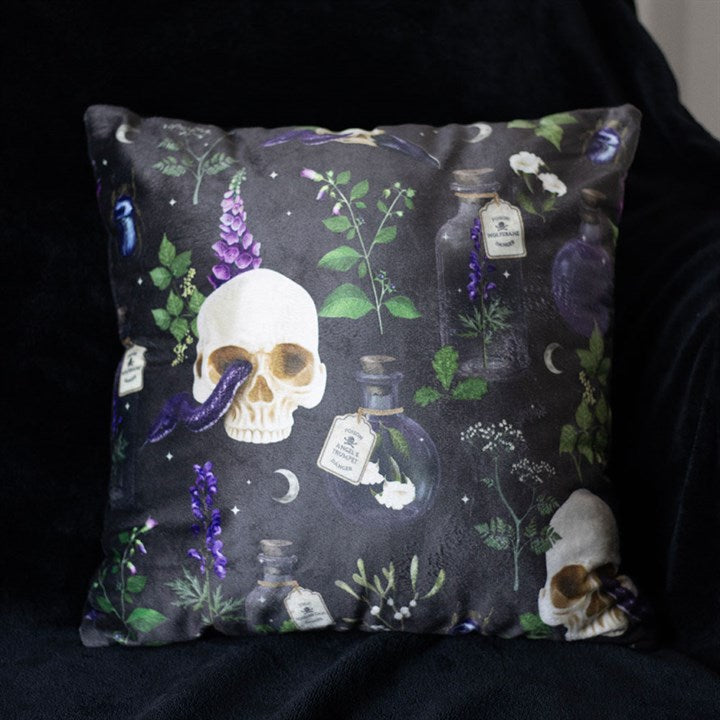 A gothic cushion with a dark background features a striking design of skulls, purple flowers, potion bottles, green leaves, crescent moons, black birds, and small white flowers.