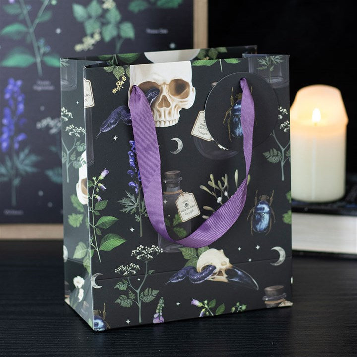 A darkly enchanting paper gift bag with a gothic twist, featuring skulls, potion bottles, moths, beetles, botanical artwork and crescent moons on black. Purple ribbon handles and a lit candle complete the look.