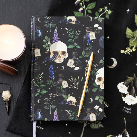 A gothic-inspired A5 paper notebook with a dark cover featuring skulls, ravens, and potion bottles rests on a black cloth; perfect for stationery enthusiasts, with a gold pen, candle, and bone charm completing the scene.