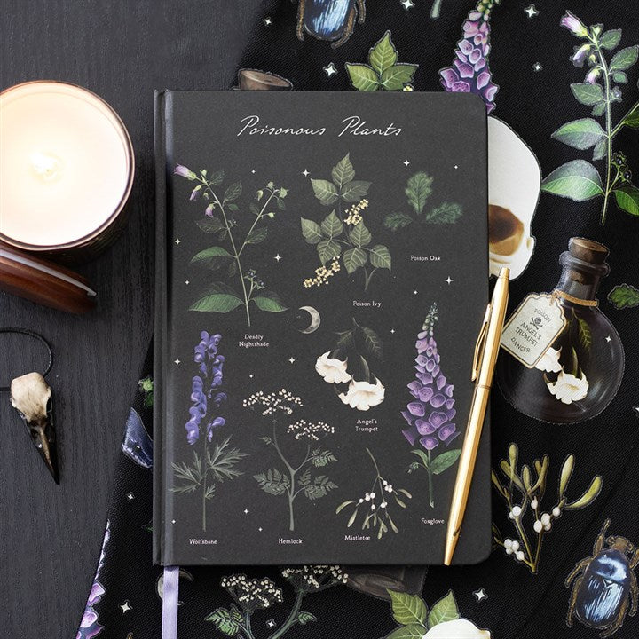 A journal titled Poisonous Plants, perfect for writing in, with illustrated plants on the A5 paper notebook cover, lies on a table alongside a candle, gold pen, botanical skull fabric, skull pendant, and decorative bottle.