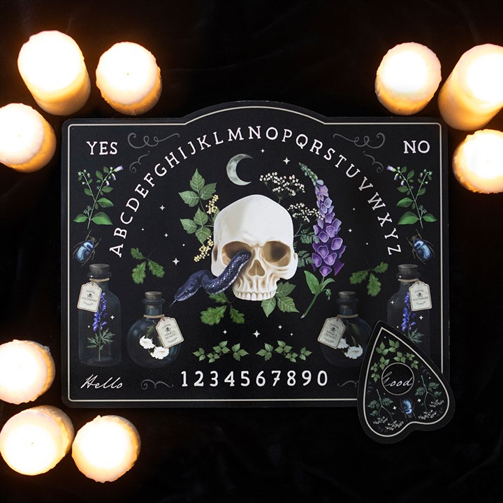 A decorative talking board featuring a central skull, flowers, potions, and a crow feather amid burning candles on black fabric. Letters, numbers, YES/NO options, and a planchette make this a truly striking decorative statement piece.