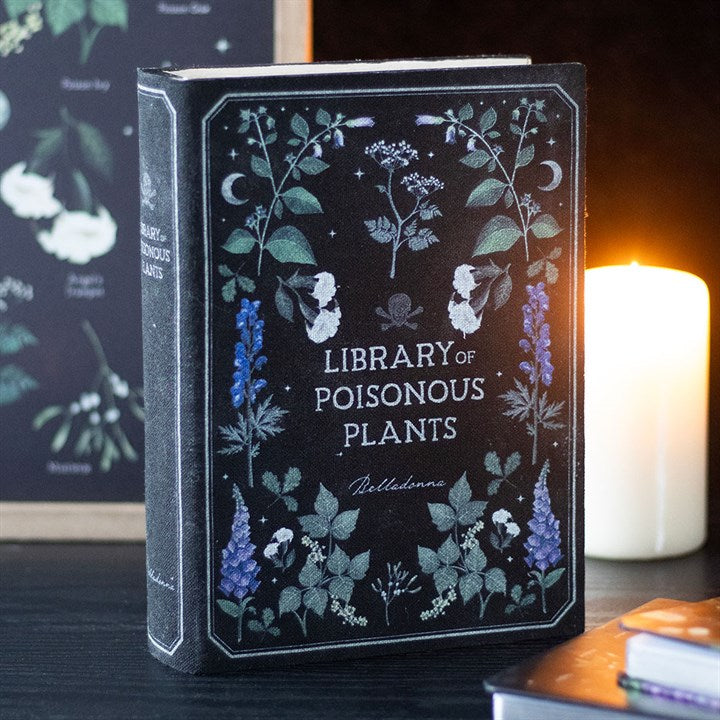 A book titled Library of Poisonous Plants stands upright on a dark table, its cover featuring botanical illustrations. A lit candle glows in the background, making it a striking addition to the home for plant or book lovers.