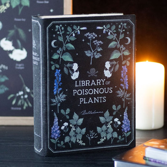 A book titled Library of Poisonous Plants stands upright on a dark table, its cover featuring botanical illustrations. A lit candle glows in the background, making it a striking addition to the home for plant or book lovers.