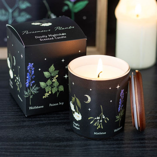 A lit Deadly Nightshade-scented candle in a black glass jar sits beside its matching box. Both feature gothic, gift-inspired botanical illustrations, stars, and a crescent moon, with the jar topped by a wooden lid.