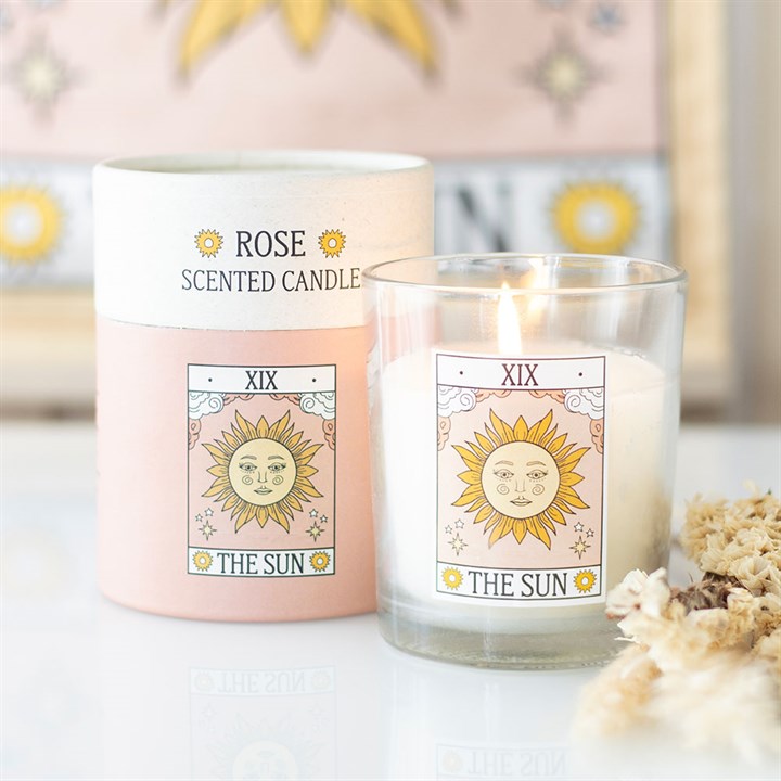 A lit rose-scented tarot card candle in a clear glass holder sits beside its pink and cream packaging, both adorned with The Sun tarot card illustration. Dried flowers add to the mystical décor.