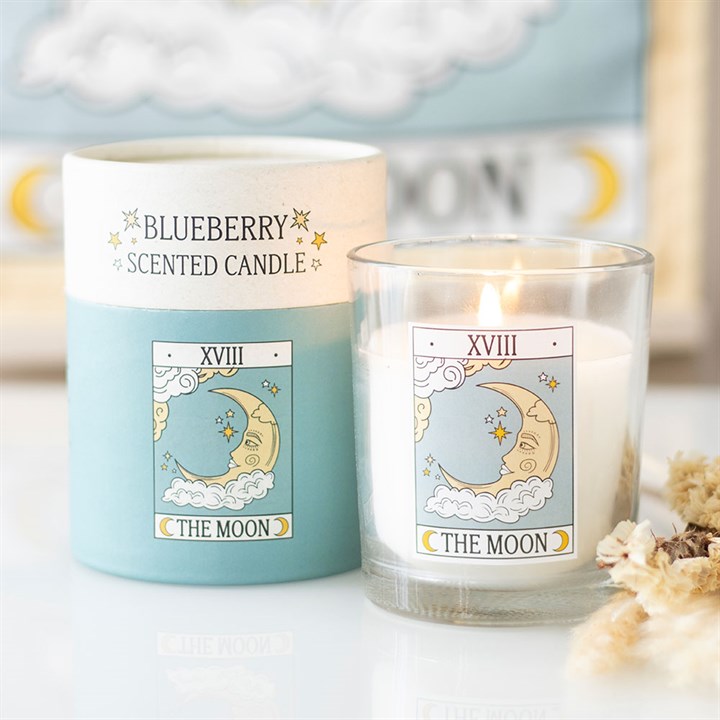 A blueberry tarot card candle in a glass sits beside its blue and white packaging, both featuring a crescent moon, clouds, and stars labelled XVIII and THE MOON. Some dried flowers add to the mystical décor.