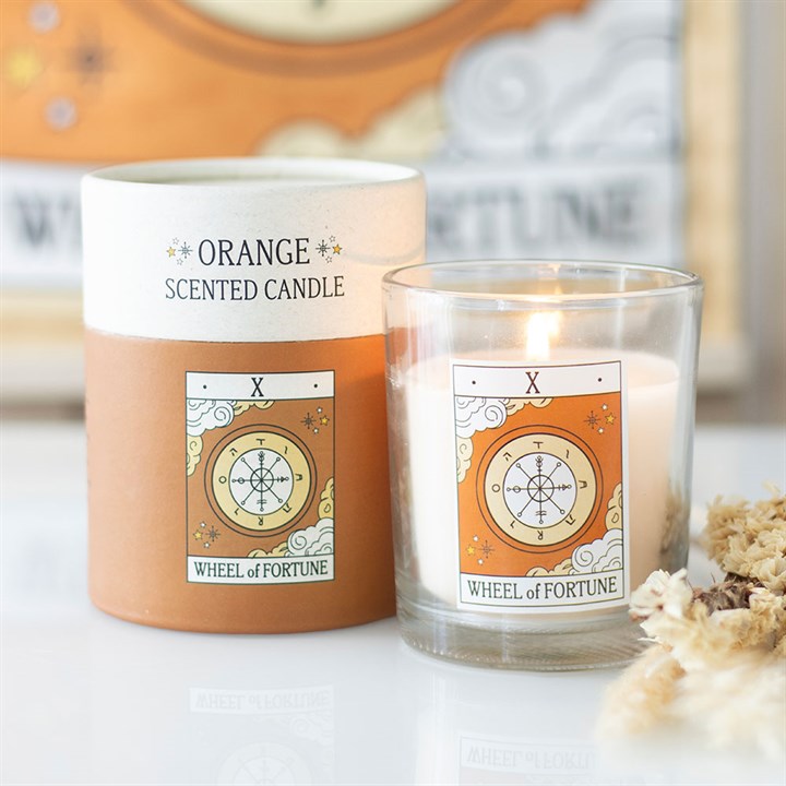 A white and orange tarot candle labelled Orange Scented Candle sits in a glass jar beside its matching box. Both feature a Wheel of Fortune design with the Roman numeral X above it. Dried flowers are nearby.