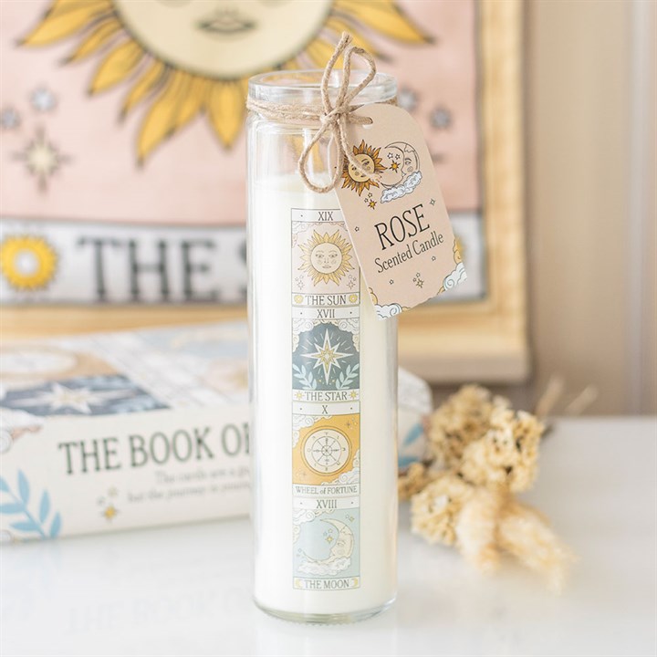 A tall tarot card candle with a Rose Scented Candle tag sits on a white surface, surrounded by dried flowers, a tarot book, and mystical décor, including a sun-themed tapestry in the background.