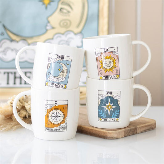 Four white tarot card mugs—The Moon, The Sun, Wheel of Fortune, and The Star—are stacked on a wooden board. This mystical mug set makes a perfect celestial gift for any tarot enthusiast. A framed moon artwork glows behind.