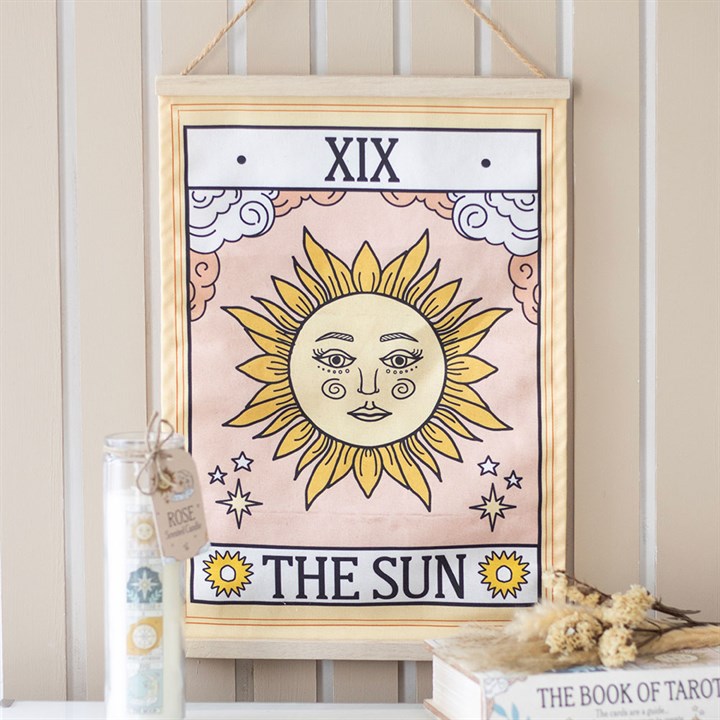 A framed Sun tarot card print with a celestial design depicts a stylised sun with a face, radiating yellow rays and pink clouds, hanging above a shelf on a striped wall.