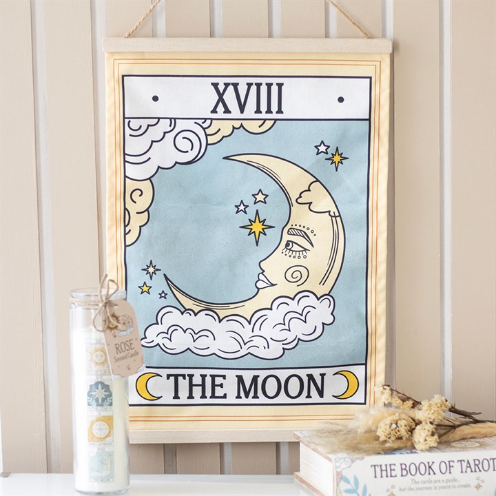 A fabric wall hanging displays “XVIII” and “THE MOON” with a celestial design—crescent moon, clouds, and stars. A floral candle and dried flowers rest beside a book titled “The Book of Tarot”.