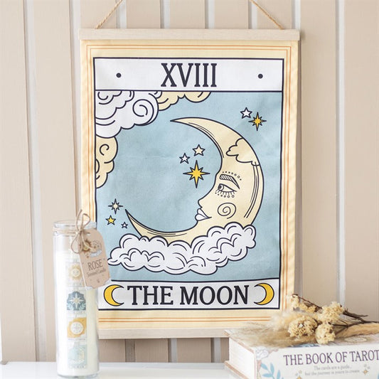 A fabric wall hanging displays “XVIII” and “THE MOON” with a celestial design—crescent moon, clouds, and stars. A floral candle and dried flowers rest beside a book titled “The Book of Tarot”.