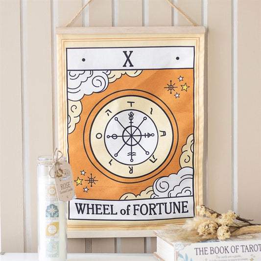 A fabric wall hanging of the Wheel of Fortune tarot card features a celestial design with a bold orange wheel, symbols, clouds, and stars above a shelf adorned with a candle, dried flowers, and a tarot book.