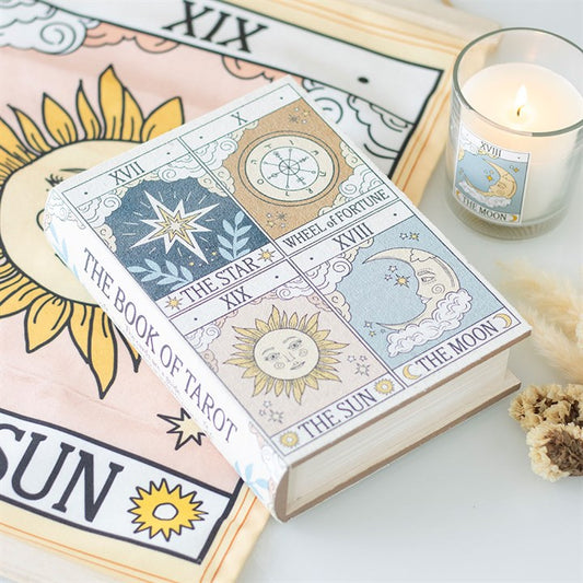 A book titled “The Book of Tarot,” with tarot card illustrations on the cover, rests on a pastel cloth featuring a celestial design. Nearby are a candle labelled ‘The Moon’, some dried flowers, and a matching tarot box.
