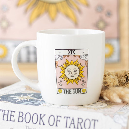 A white Sun tarot mug features an illustration of the Sun tarot card (XIX), perfect for tarot enthusiasts. It rests on The Book of Tarot with dried flowers nearby, adding a mystical touch to your space.
