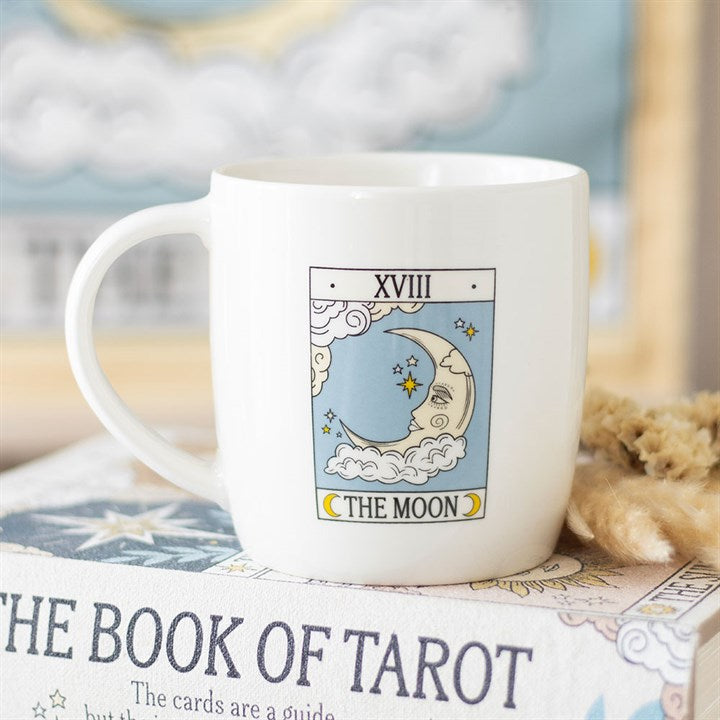 A white The Moon tarot mug features an illustration of the card with a crescent moon, stars, clouds, and XVIII above. Perfect for tarot enthusiasts, the mug rests on a book titled The Book of Tarot. Rewritten in British English: A white The Moon tarot mug features an illustration of the card with a crescent moon, stars, clouds and XVIII above. Perfect for tarot enthusiasts, the mug is placed on a book entitled *The Book of Tarot*.