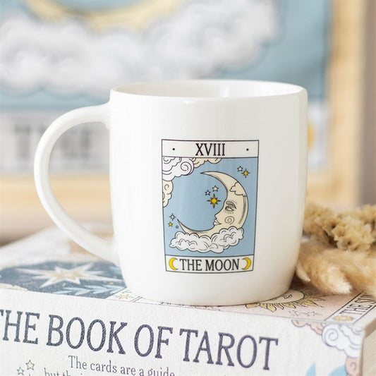 A white The Moon tarot mug features an illustration of the card with a crescent moon, stars, clouds, and XVIII above. Perfect for tarot enthusiasts, the mug rests on a book titled The Book of Tarot. Rewritten in British English: A white The Moon tarot mug features an illustration of the card with a crescent moon, stars, clouds and XVIII above. Perfect for tarot enthusiasts, the mug is placed on a book entitled *The Book of Tarot*.