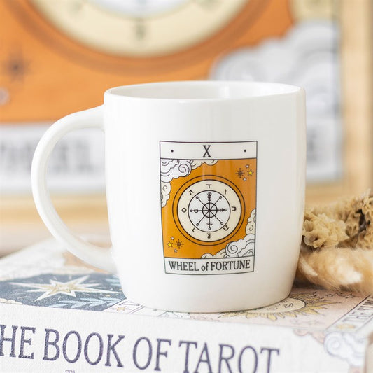 A white tarot mug rests on a tarot book, featuring the Wheel of Fortune card design with its iconic wheel, symbols, and cloud motifs in gold, white, and black—perfect for tarot enthusiasts.