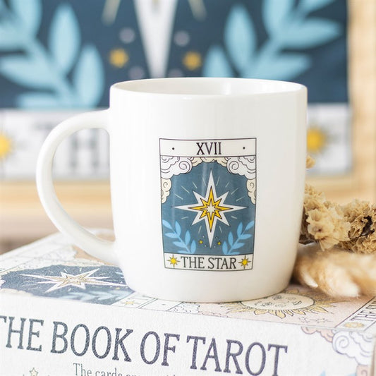 A celestial white The Star tarot mug featuring XVII THE STAR with a yellow star illustration rests atop THE BOOK OF TAROT, making it a perfect find for tarot enthusiasts. Dried flowers complete the scene. Rewritten in British English: A celestial white The Star tarot mug, featuring XVII THE STAR with a yellow star illustration, rests atop THE BOOK OF TAROT, making it a perfect find for tarot enthusiasts. Dried flowers complete the arrangement.