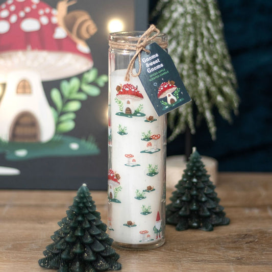 A white Gnome Sweet Gnome candle in a glass jar rests on a wooden surface, adorned with red-capped mushroom houses and gnomes. It is surrounded by small green winter forest candles shaped like trees.