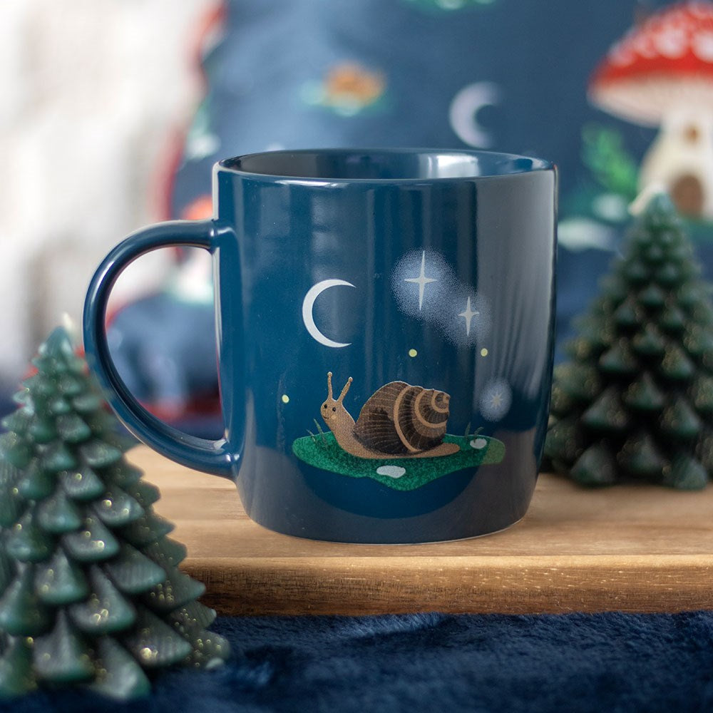 A navy-blue mug featuring a whimsical snail on grass, surrounded by stars, a crescent moon and sparkles. The mug sits on a wooden tray with two pine tree candles. A mushroom cushion is gently blurred in the background.