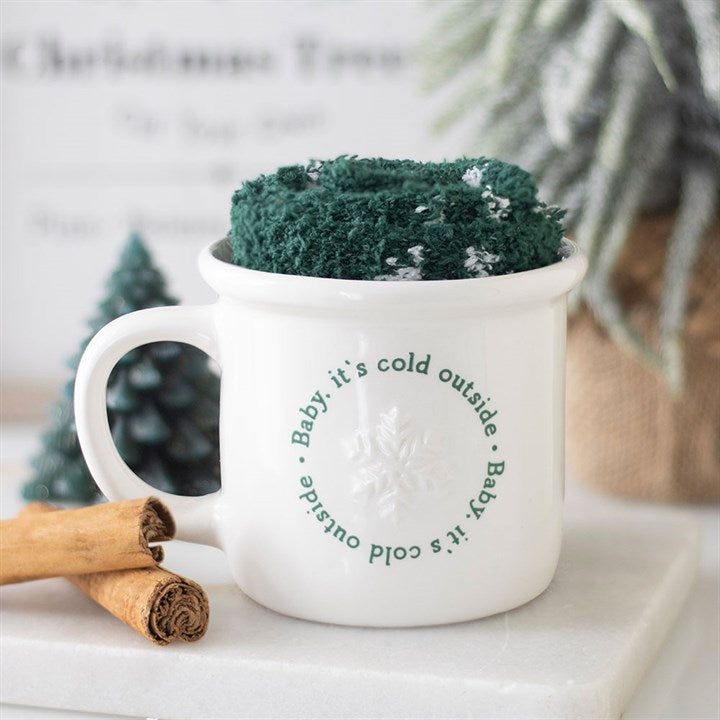 A festive ceramic mug with Baby, it’s cold outside in green lettering and a snowflake design holds cosy socks. Two cinnamon sticks and small pine tree decorations sit nearby on a white surface.