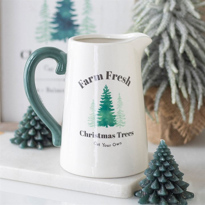 A white ceramic flower jug with a green handle features the text “Farm Fresh Christmas Trees Cut Your Own” and illustrations of trees. Perfect for farmhouse-style Christmas décor, it sits amongst festive candles and faux pine branches.