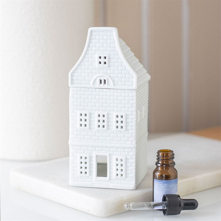 A small white ceramic oil burner shaped like a Nordic house, featuring textured brick details, sits on a white surface next to a brown glass dropper bottle against a blurred beige background.