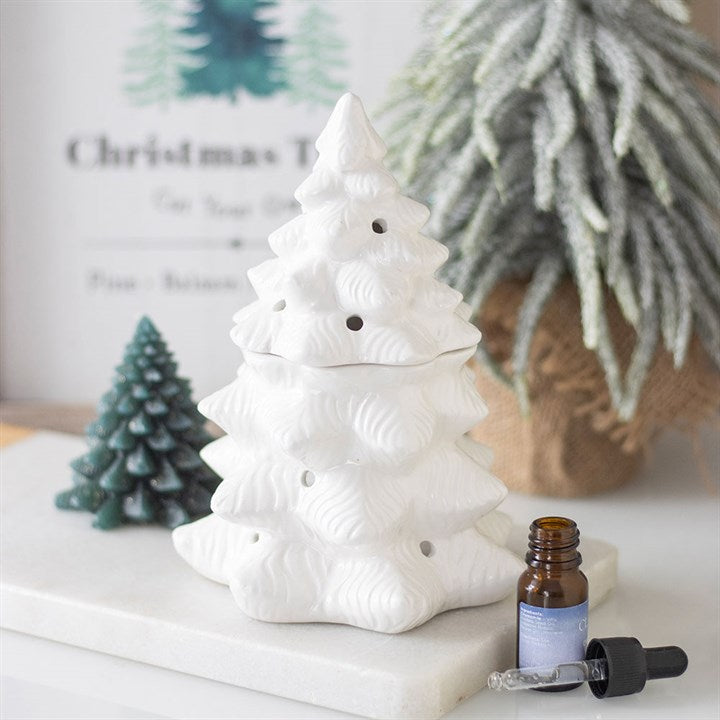 A white ceramic Christmas tree oil burner sits on a white tray, with an amber essential oil bottle, a dropper, and a green tree-shaped candle nearby; pine tree décor is blurred in the background.
