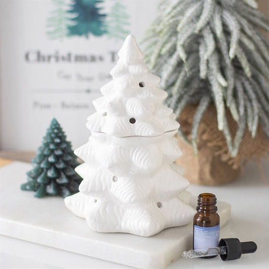 A white ceramic Christmas tree oil burner sits on a white tray, with an amber essential oil bottle, a dropper, and a green tree-shaped candle nearby; pine tree décor is blurred in the background.