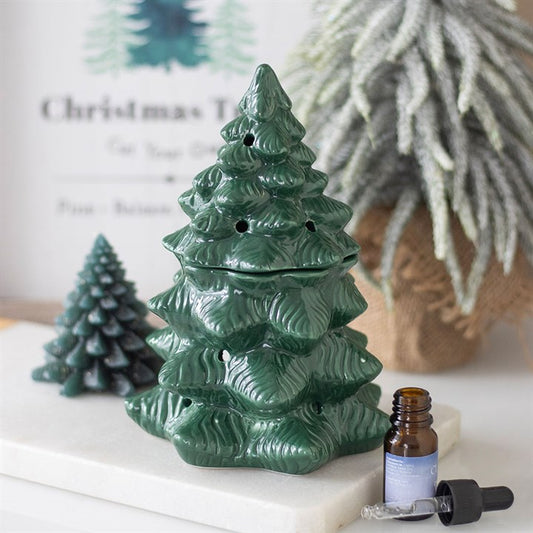 A glossy green ceramic oil burner shaped like a white fir tree sits on a white surface, accompanied by an open brown essential oil bottle, dropper, and a smaller tree ornament. Blurred festive decorations appear in the background.