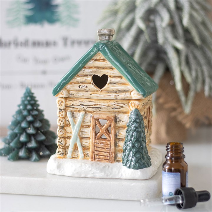 A ceramic ski chalet oil burner with a green roof and heart-shaped window sits on artificial snow, flanked by pine trees. Nearby are an amber dropper and an open essential oil bottle, with a blurred Christmas sign in the background.