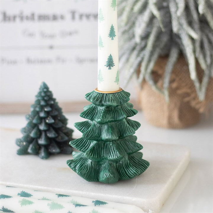 A green, Christmas tree-shaped candle holder holds a white candle adorned with small green fir trees. A smaller tree decoration and a frosted plant in hessian sit blurred in the background on a white surface.