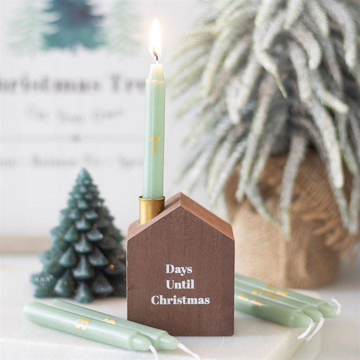 A lit green Advent candle stands in a brown wooden, house-shaped holder labelled Days Until Christmas. Festive decorations and spare green candles are in the background on a light-coloured surface.