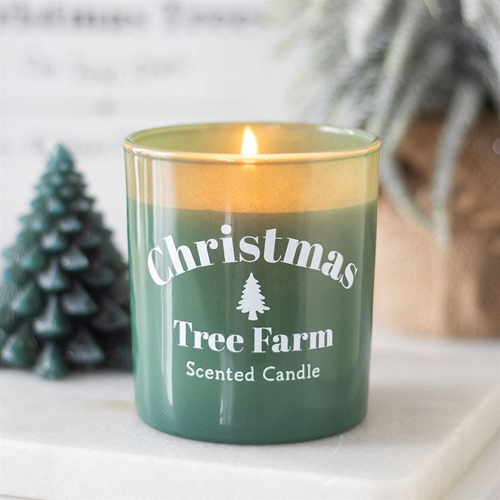 A green Christmas Tree Farm candle burns with a gentle flame. A small tree-shaped decoration and a blurred plant in the background create a festive, cosy atmosphere. Perfect as a festive candle for winter evenings.