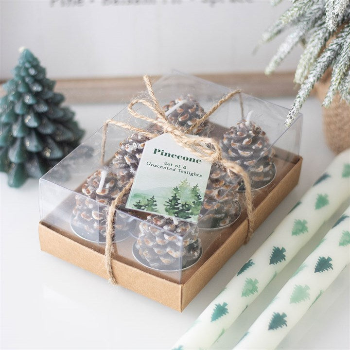 A clear box tied with string holds six pinecone tealight candles. The label reads ‘Pinecone Set of 6’. These Christmas candles are perfect for woodland décor, with tree-shaped and patterned white candles nearby.