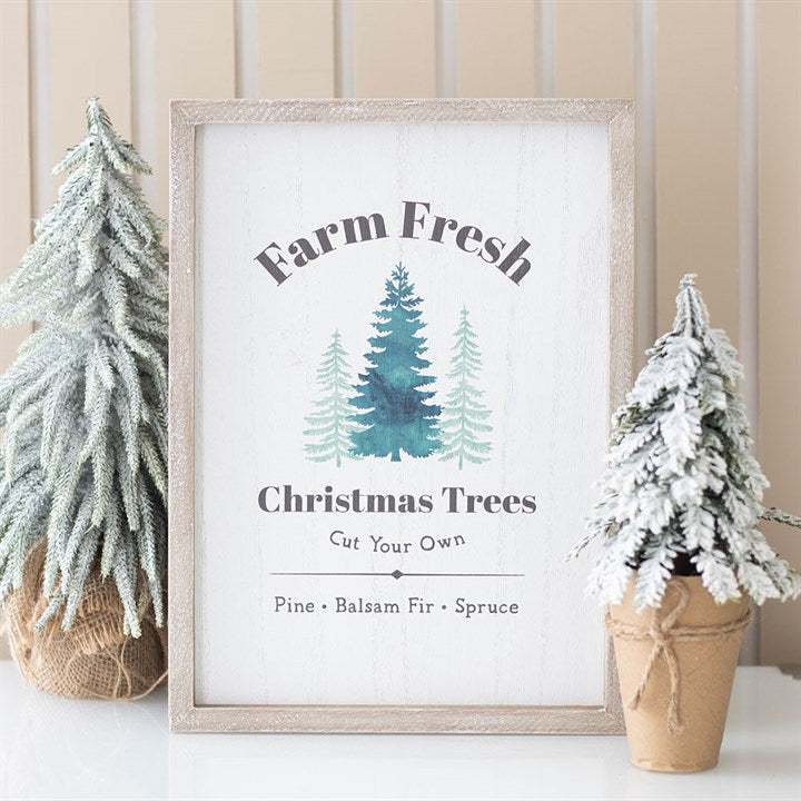 A framed sign reads ‘Farm Fresh Christmas Trees Cut Your Own’, perfect for festive décor, featuring painted pine trees and snow-dusted potted evergreens set against a light wood-panelled, farmhouse-style background.