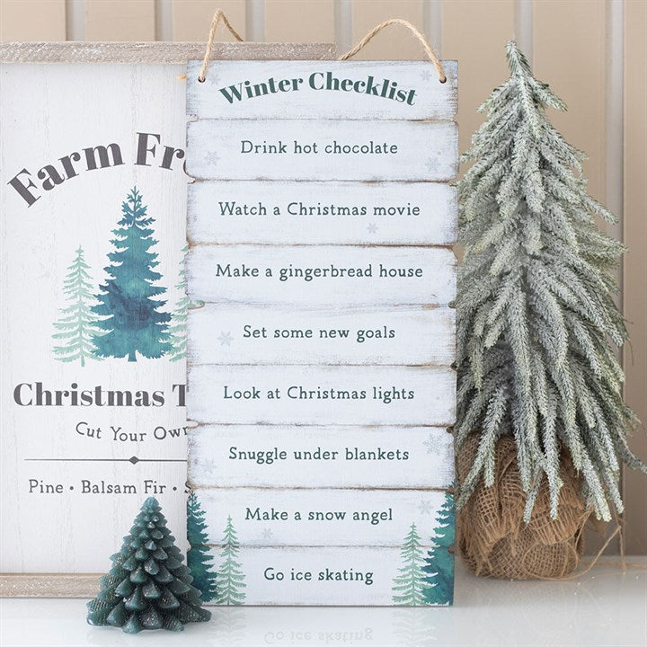 A wooden Winter Wonderland Checklist sign lists festive activities such as drinking hot chocolate and making a gingerbread house. It sits between two small artificial pine trees, perfect as Christmas décor.