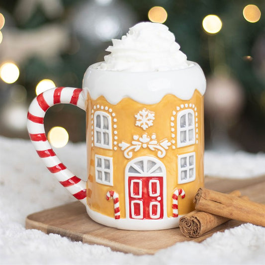 A festive Christmas mug shaped like a gingerbread house, topped with whipped cream, sits beside two cinnamon sticks on a wooden board; blurred lights glow in the background.
