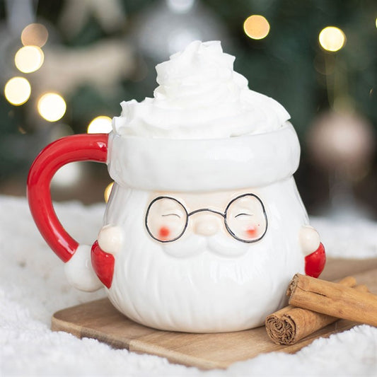A Father Christmas mug shaped like his smiling face is filled with whipped cream, capturing the Christmas spirit. The mug’s red handle, black spectacles and rosy cheeks glow amidst blurred festive lights and greenery.