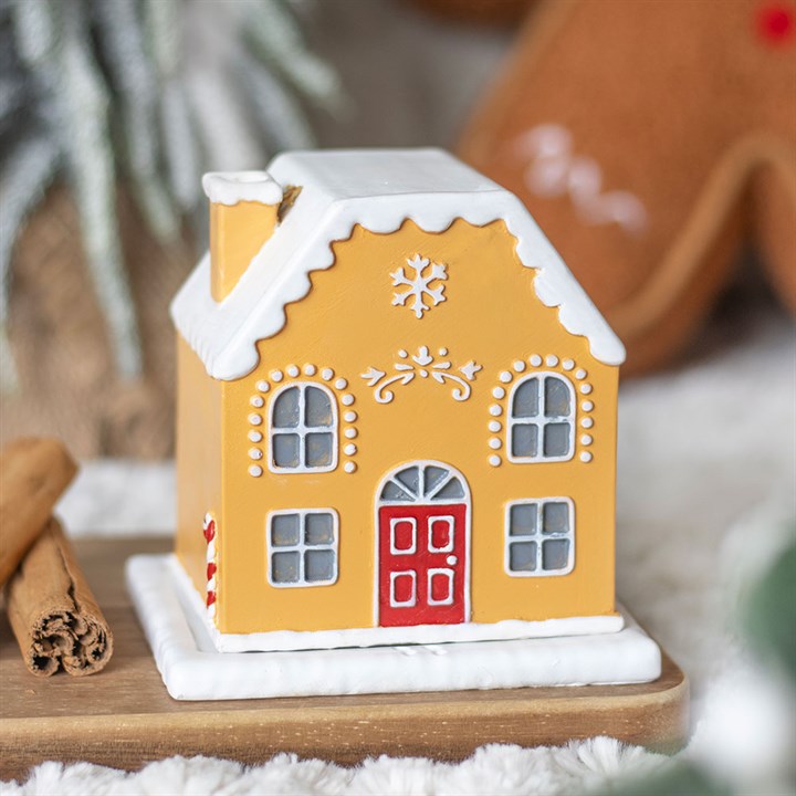 A small, yellow gingerbread house incense holder with white icing details, a red door, and a snowflake design sits on a wooden tray next to two cinnamon sticks; perfect for festive home décor.