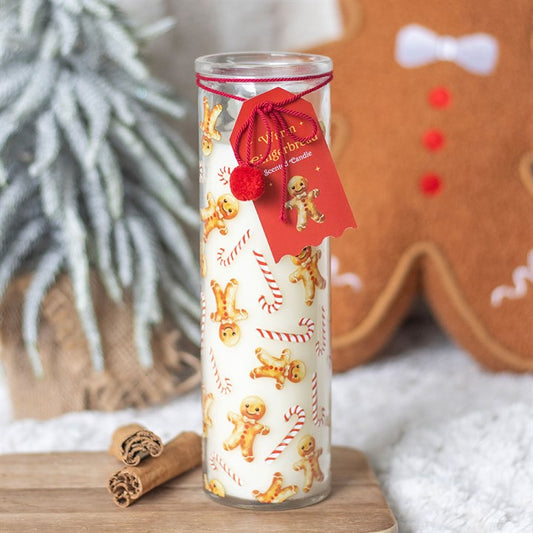 A festive candle decorated with gingerbread men and candy canes sits on a wooden surface. Cinnamon sticks lie beside it, with a gingerbread figure and a pine branch in the softly blurred background.