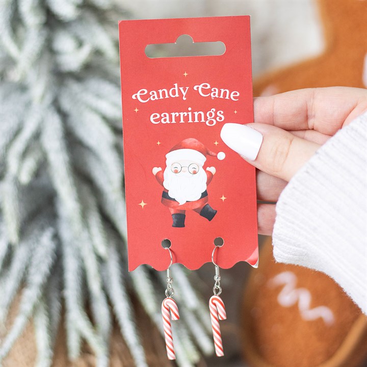 A hand with white polished nails holds a red card labelled Candy Cane Earrings, featuring a cartoon Santa. Two dangling Christmas earrings shaped like red and white candy canes are attached, with a blurred festive background.