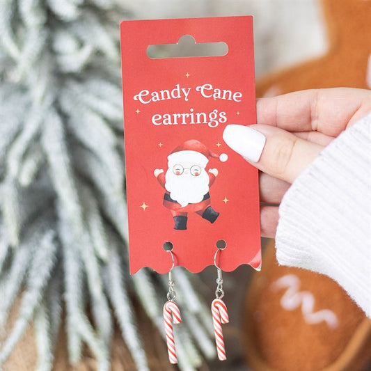 A hand with white polished nails holds a red card labelled Candy Cane Earrings, featuring a cartoon Santa. Two dangling Christmas earrings shaped like red and white candy canes are attached, with a blurred festive background.