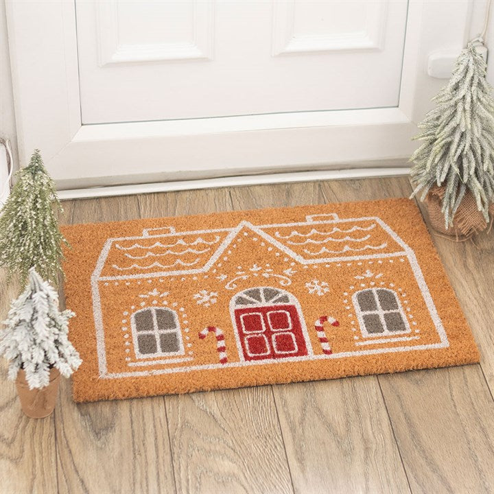 Channelling the seasonal spirit, this festive Gingerbread House doormat, featuring red doors, grey windows, candy canes, and snowflakes, sits before a white door and is flanked by snow-dusted pine trees on a wooden floor.