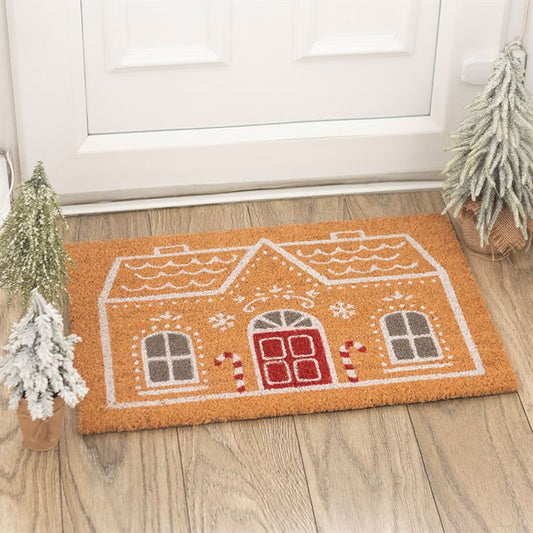 Channelling the seasonal spirit, this festive Gingerbread House doormat, featuring red doors, grey windows, candy canes, and snowflakes, sits before a white door and is flanked by snow-dusted pine trees on a wooden floor.