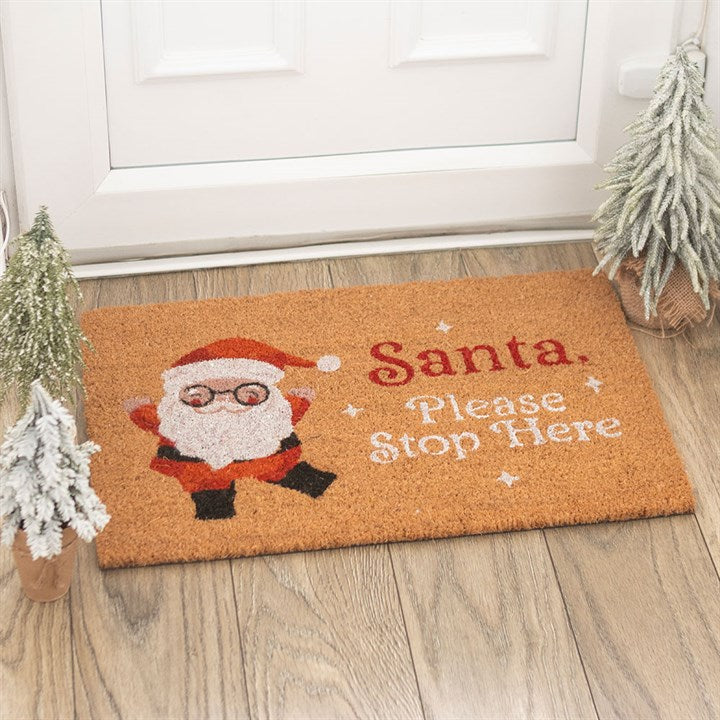 A festive Santa Stop Here doormat featuring a cartoon Father Christmas waving and bold red text brightens the wooden doorway, with two frosted artificial trees flanking this cheerful Christmas doormat.