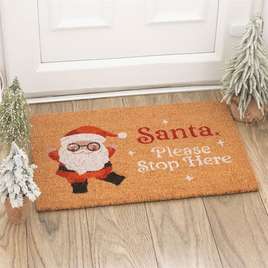A festive Santa Stop Here doormat featuring a cartoon Father Christmas waving and bold red text brightens the wooden doorway, with two frosted artificial trees flanking this cheerful Christmas doormat.