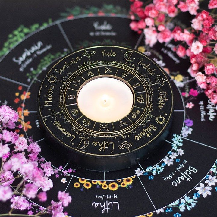 A black resin tealight holder, adorned with the names of Wiccan sabbats in gold, encircles a lit tealight and rests upon a colourful Wheel of the Year chart. Clusters of pink and purple flowers add accents to the scene.